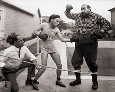 Boxeando con el actor Mack Swain con su vestuario de La quimera del oro (The Gold Rush). El árbitro es el boxeador real Kid McCoy (1923-1925)