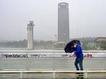 Un persona se protege de la lluvia y el viento caminando por la pasarela peatonal de la Cartuja en Sevilla.