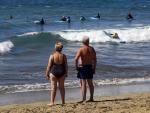 Una pareja de turistas observa c&oacute;mo varios j&oacute;venes toman clases de surf este jueves santo en la playa de Maspalomas, en Canarias.