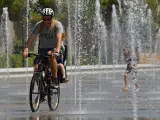 Un padre y su hijo en bicicleta pasan por las fuentes del Parque Central de Valencia mientras una ni&ntilde;a juega con el agua en el fondo.