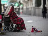 Una persona en silla de ruedas y tapada con una manta pide dinero con un muñeco