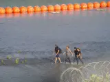 Migrantes caminan entre alambre de concertina y una cadena de boyas colocadas en el agua a lo largo de la frontera del Río Grande con México en Eagle Pass, Texas.