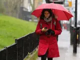 Una mujer consulta tu teléfono móvil mientras camina bajo la lluvia con un paraguas.