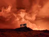 Varias personas observan desde la lejanía cómo se eleva una gran columna de humo, mientras la lava tiñe el cielo nocturno de naranja por la erupción del volcán islandés.