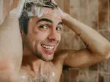 A man stands in the shower, scrubbing his head and creating a lather with the shampoo. He looks at the camera and smiles as he does so.