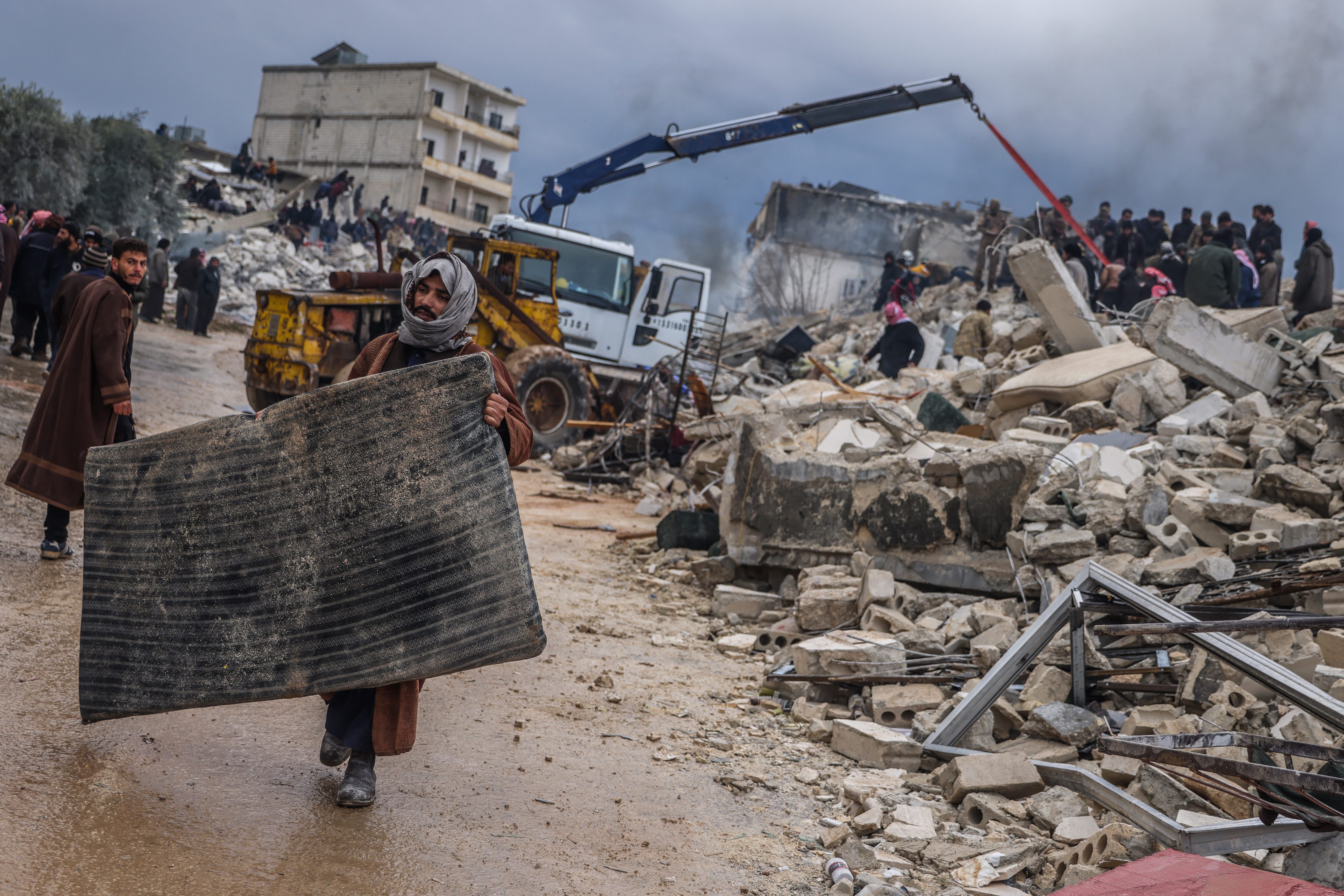Un hombre, con una esterilla para protegerse del frío, pasa junto a un edificio derruido por el terremoto, donde varias personas buscan supervivientes entre los escombros, en Harem (Siria).